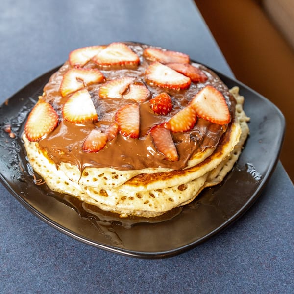 Stack of Ollie's pancakes topped with chocolate sauce and sliced strawberries on a black plate.