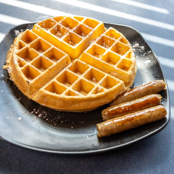 A golden waffle dusted with powdered sugar and three breakfast sausages on a black plate.